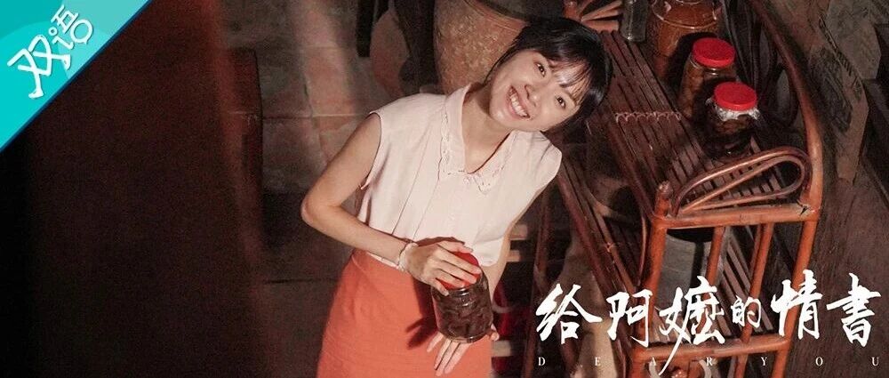 A film still from Dear You showing a young woman smiling warmly while holding a glass jar in a dim period kitchen