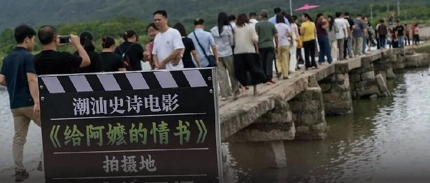 Crowds of visitors gathered at an old stone bridge by the water, beside a clapperboard sign marking it as a filming location for Dear You